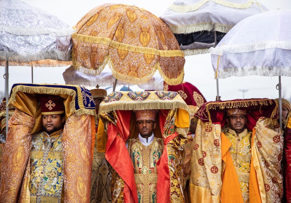 Priest wearing gold traditional garb carry covered tablets over their heads. Umbrellas fill the upper half of the frame.