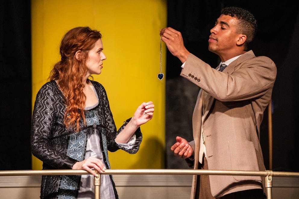 Woman, left, in blue dress and with red long curly hair stands next to a man, in a beige suit, holding up a pendant