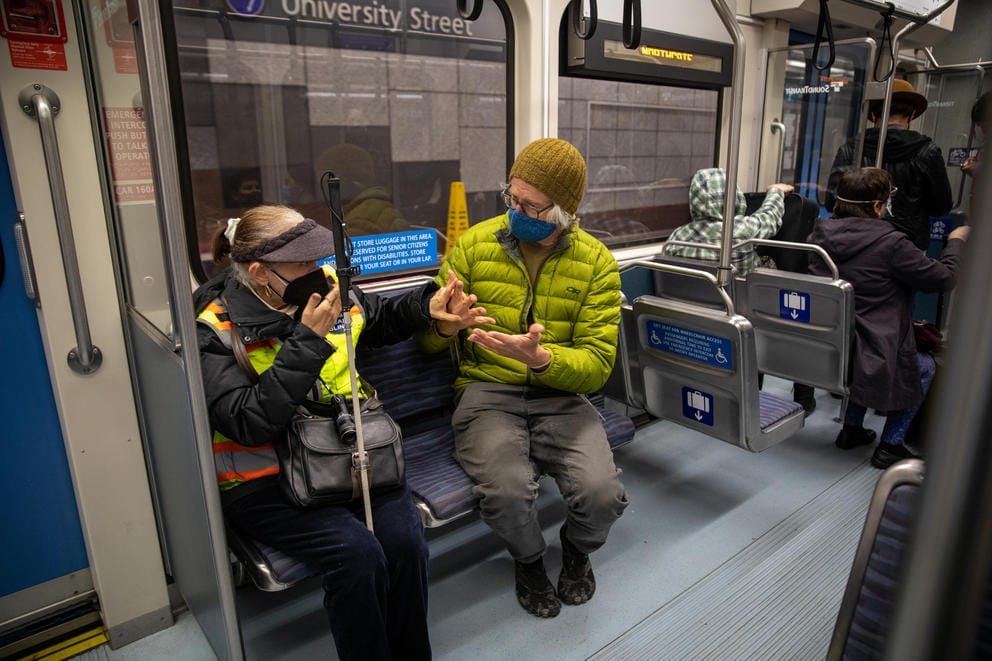 A medium shot of Lynn Chase, wearing a neon safety vest and black visor with a cane wedged between her knees, and orientation and mobility trainer David Miller, wearing a green jacket, using tactile sign language while sitting on a bench on a light rail train. Other passengers can be seen in the background.