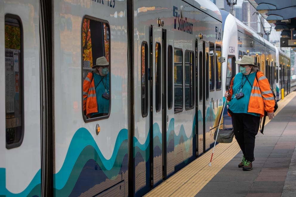 A wide shot of Dorene Cornwell in neon orange sweatshirt waiting at the Sodo Station platform as a train pulls in.