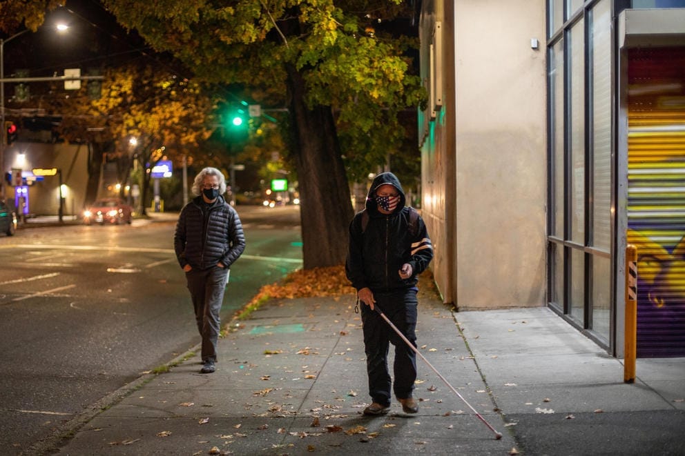 A wide shot of Chris Loomis, wearing a black sweatshirt and pants, and American flag mask, walking down a street using a cane and miniguide, while David Miller, an orientation and mobility trainer, wearing a black jacket, followed early one fall morning before the sun was up.
