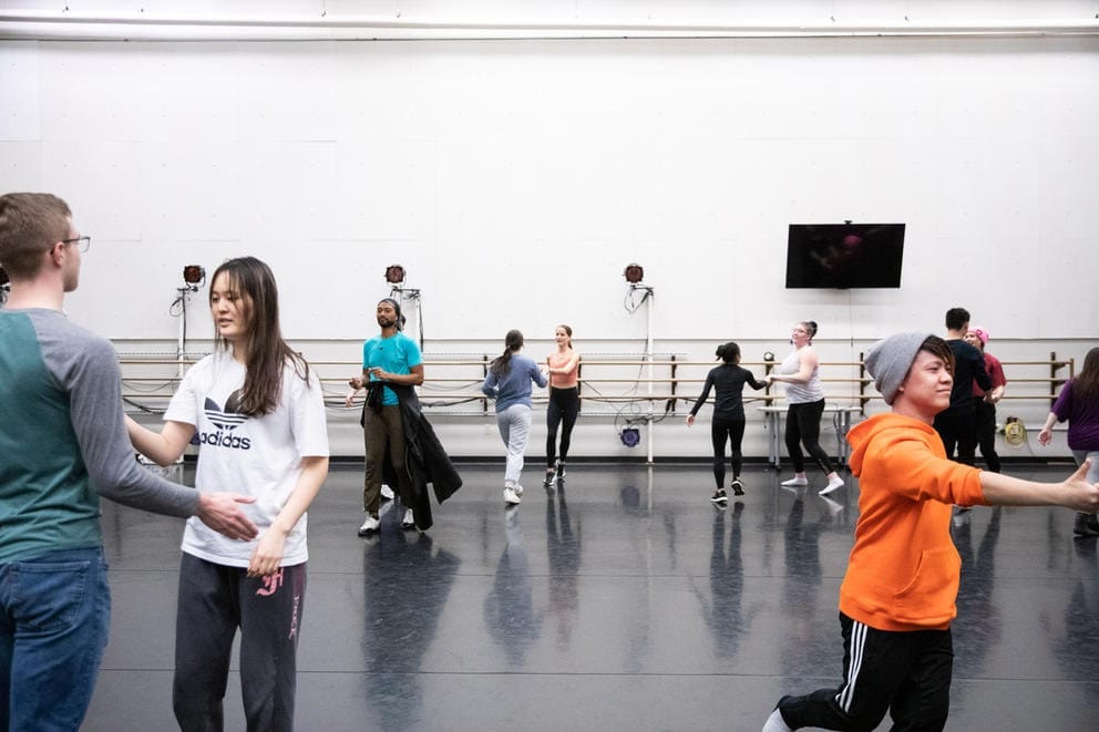 People in a rehearsal dance room with dark flooring are practicing steps in pairs