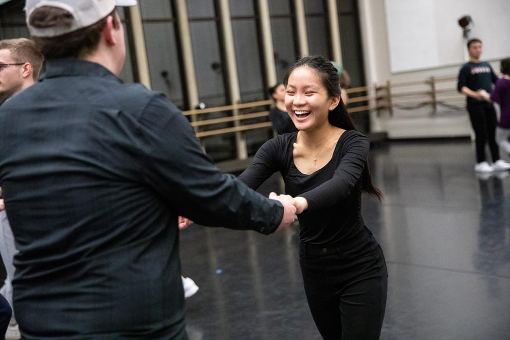 Person holds the hands of another person, both dressed in black, as they are dancing together. The person on the left is smiling widely. They are in a rehearsal space.