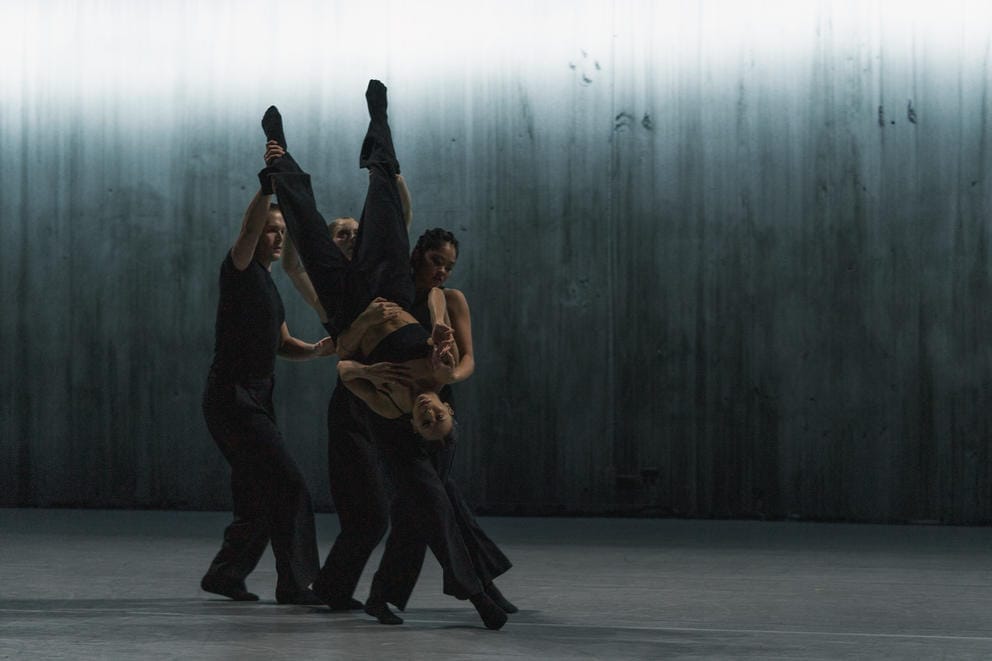 a tight group of dancers, on upside-down and carried by the others, on a dark stage