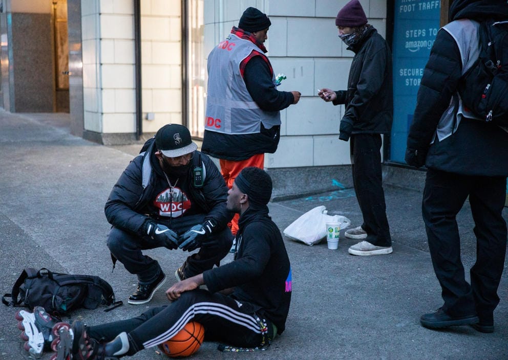 a man wearing rollerblades sits on the sidewalk while another man kneels down to offer help