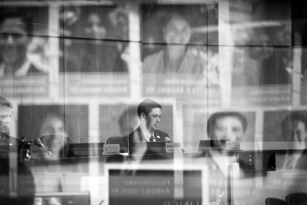 reflections of several city councilmembers in plexiglass inside city hall