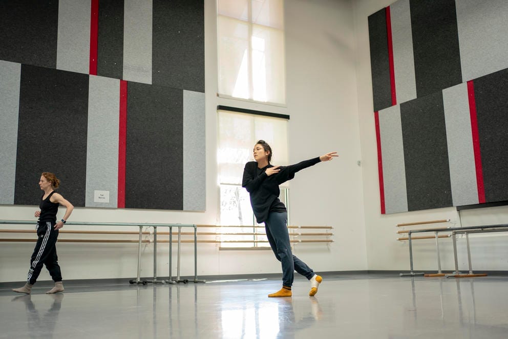 A wide shot of a dancer rehearsing in a studio, the choreographer walks out of the frame at the left