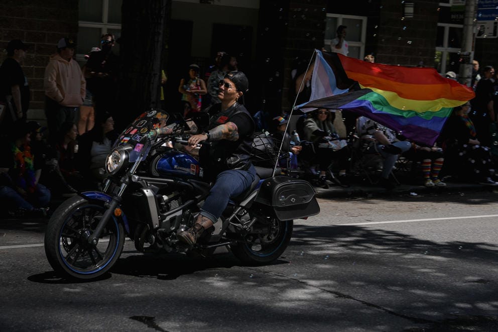 A motorcycle rider is lit up by a patch of sun. A rainbow Pride flag blows behind the bike.