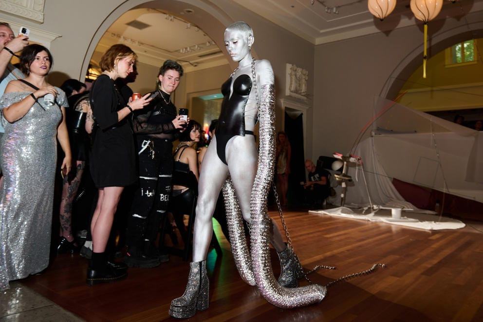 A model in silver paint and a black body suit walks down the runway; the audience takes photos
