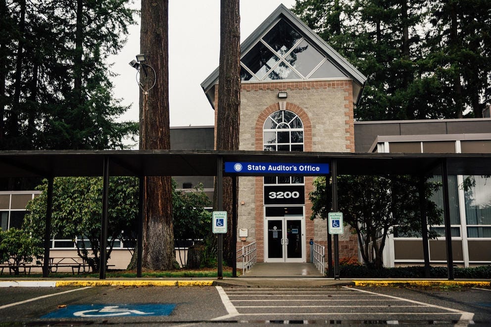 A blue sign reads State Auditor's Office over entrance to a government building in a wooded area.