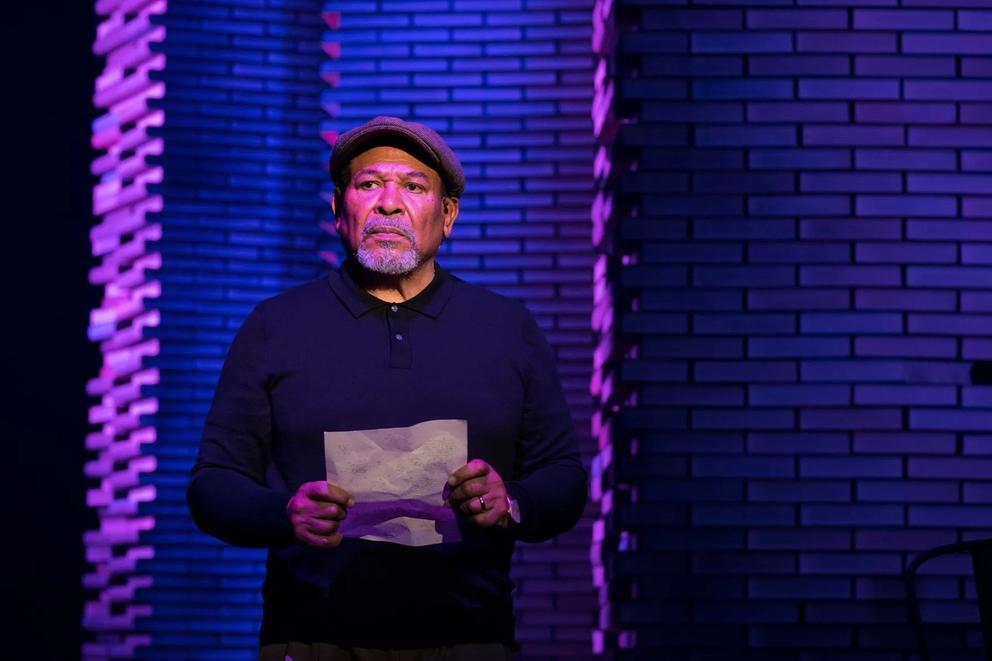 photo of an indigo-lit stage play with a single man in a hat holding a piece of paper