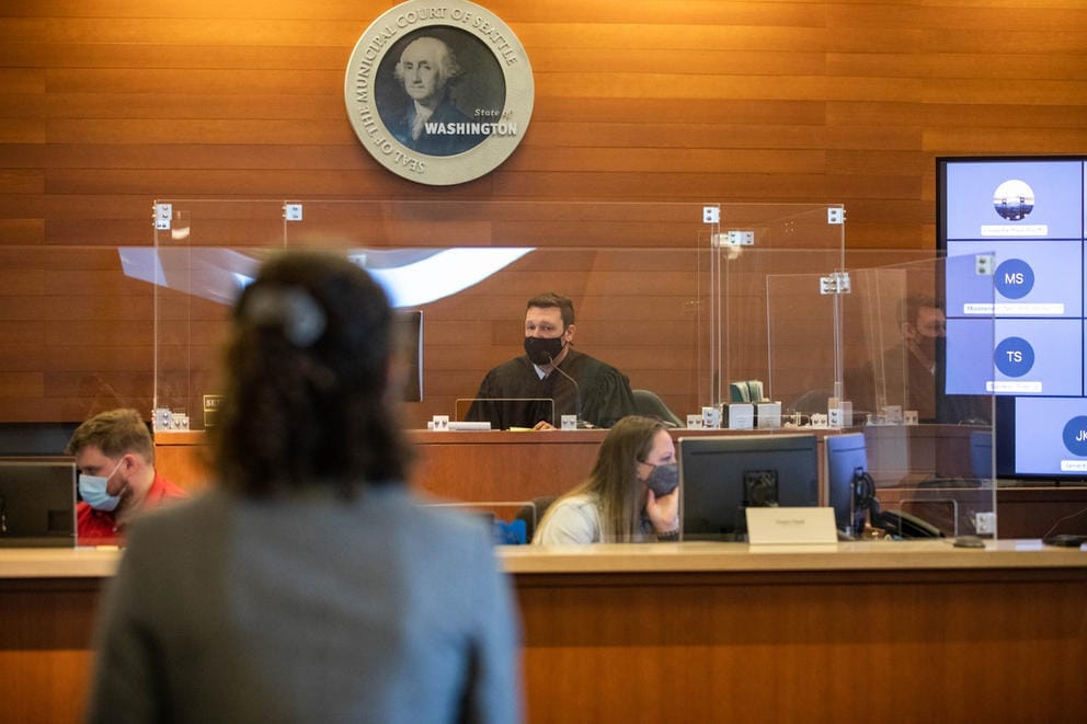 A judge seated at the front of a courtroom wearing a mask