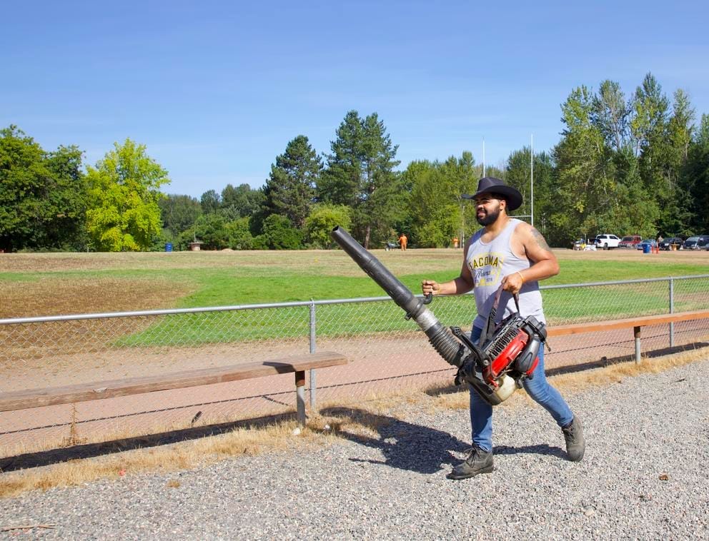 A person in a cowboy hat carries a leaf blower across a dirt path in the foreground of a ball field. There are trees in the background.