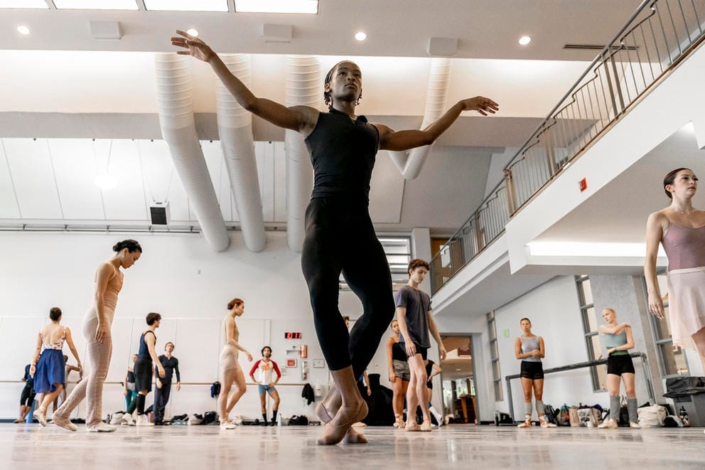 A ballerina at the front of the image outstretches his arms and plies; other dancers walk in the background
