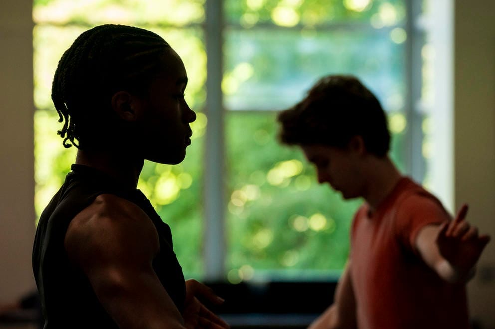 A side profile of a person looking straight ahead; behind them, another person is looking down with one arm outstretched in a dance pose