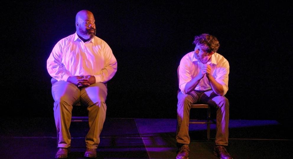 two men in business casual clothes sit in chairs on stage in a purplish light