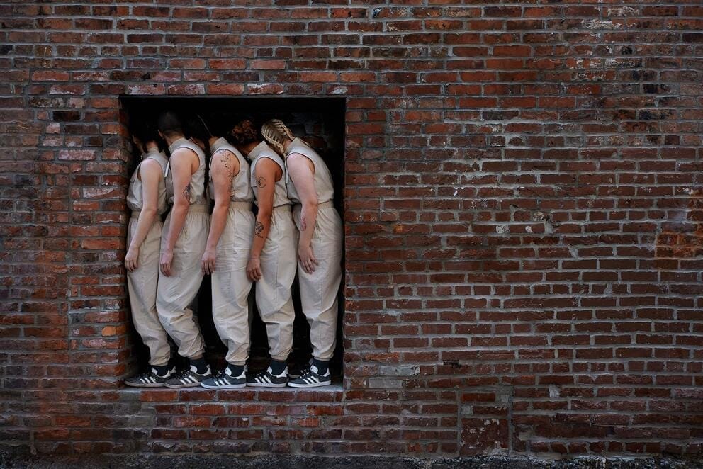 a group of dancers in tan jumpsuits gathers tightly in an opening in a brick wall