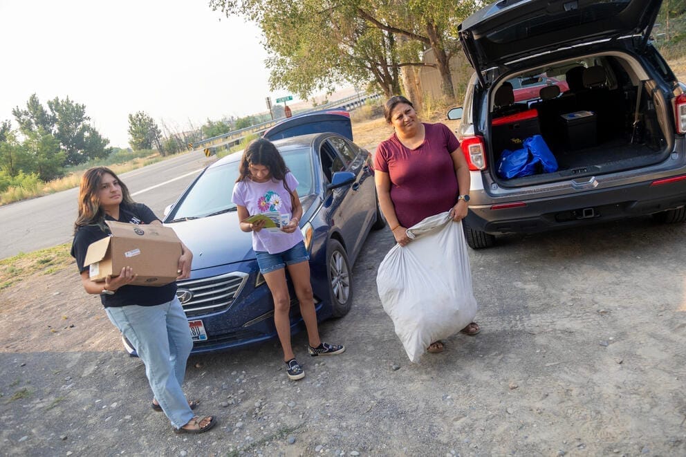Three people, one holding a bag and another box, walk away from cars parked in a field.