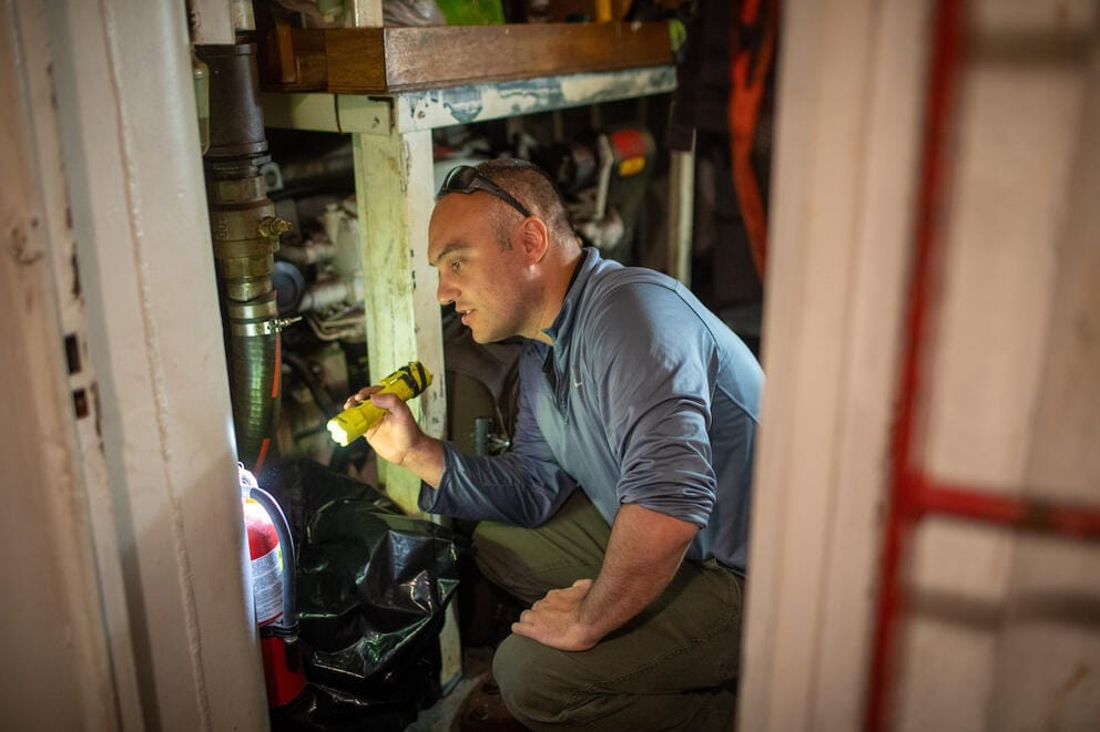 Cyrus Donato inspects below deck on a ship with a flashlight in hand.