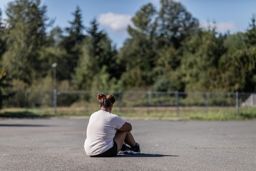 A person with their back turned to the camera sits on an asphalt surface near a fenced off wooded area.
