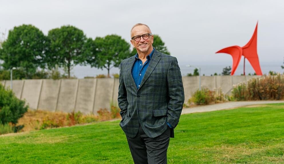 a smiling man in glasses stands outdoors in a sculpture park
