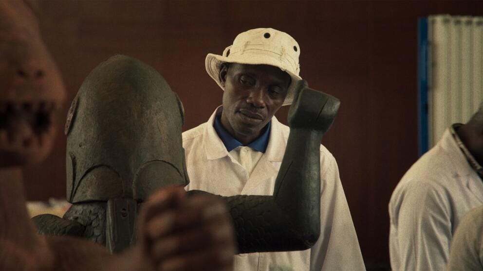 still from a film with a man in a hat looking at a Benin sculpture