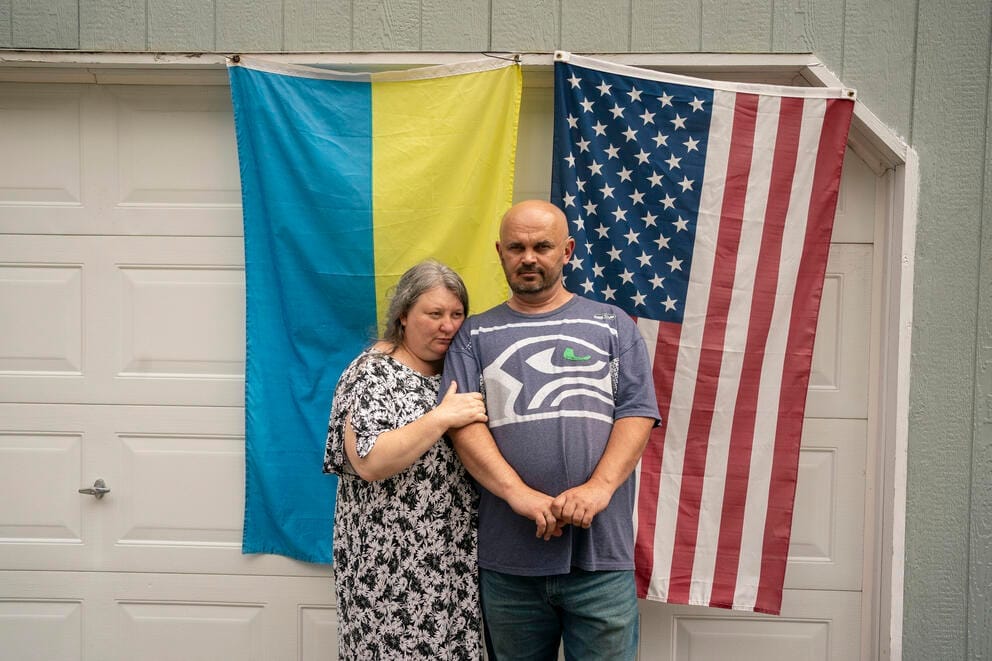 A woman and man stand in front of a garage with a Ukrainian flag and an American flag hanging behind them