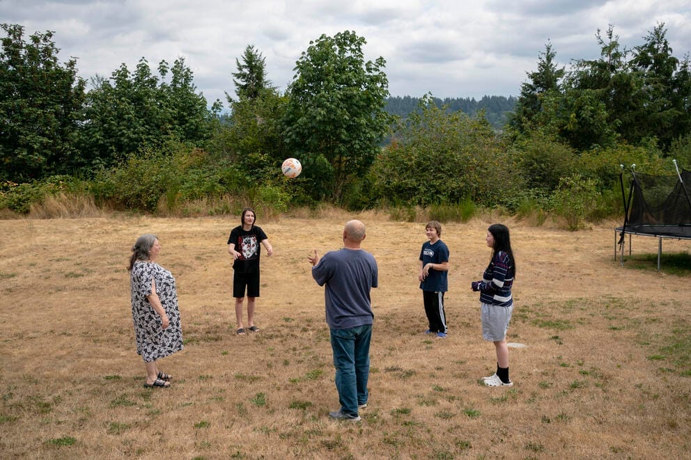 A group of five people, including parents and their three children stand in a circle in a field tossing a soccer ball in the air
