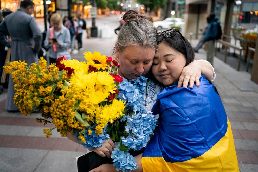 Two women hold each other on a city street, the one on the left is holding a bouquet of yellow flowers, the one on the right has a Ukrainian flag draped around her shoulders.