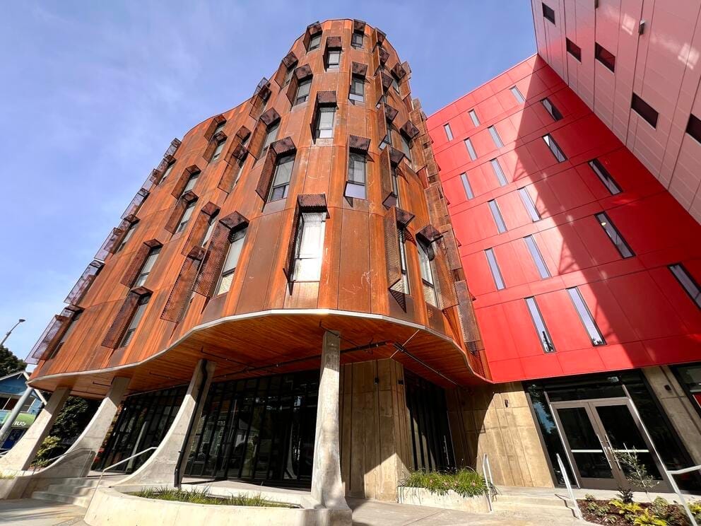 exterior of a windowed building with a curving brown facade set on arced supports, next to a red rectilinear building
