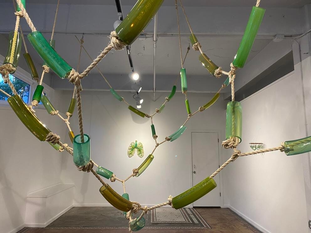 a white walled gallery is hung with hanging ropes partially encased in tubes made of green glass