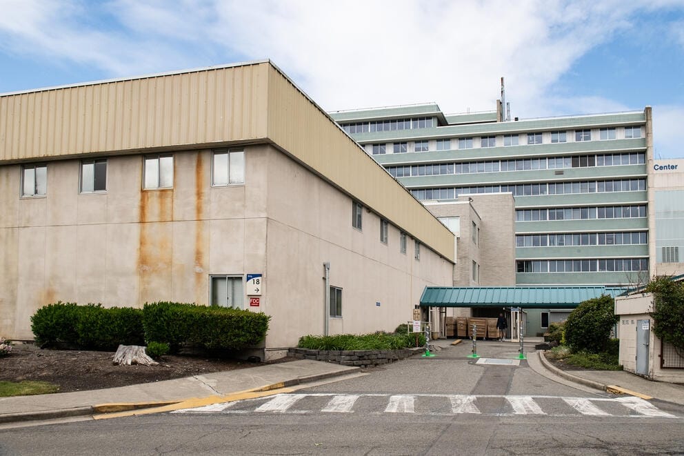 A beige building sits in front of a greenish building lined with windows.
