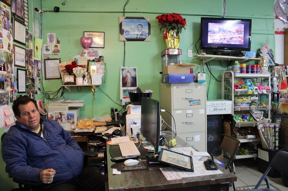 A person sits in an office with a variety of greeting cards, flowers, and other certificates and pictures on the wall.