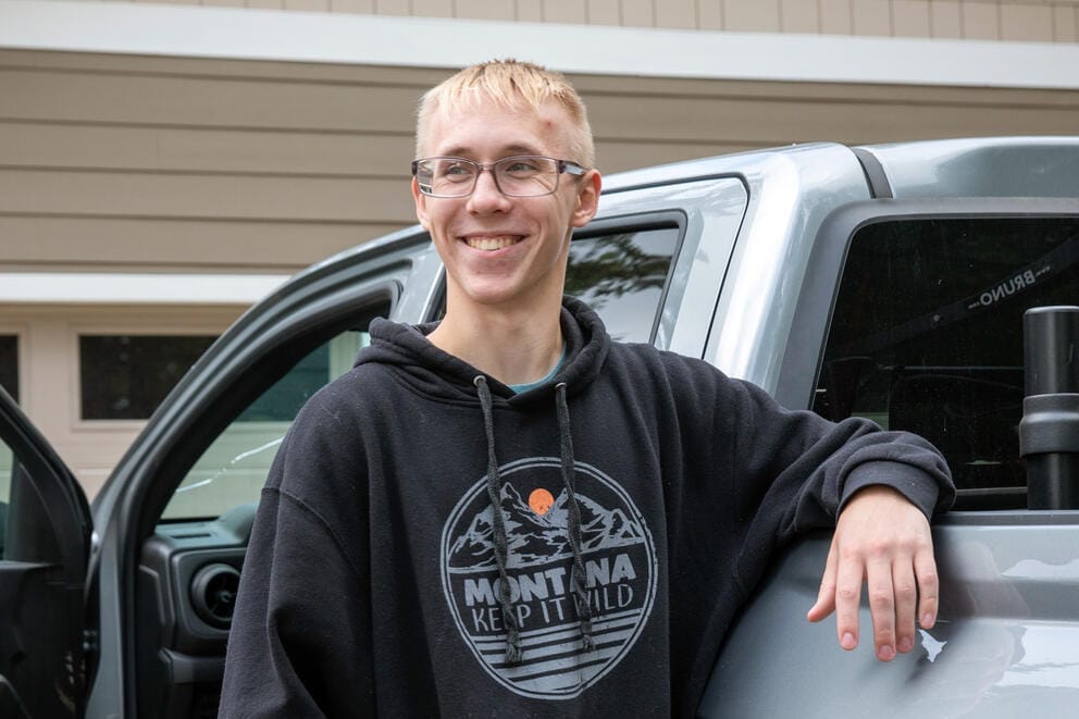 A portrait of a person with blond hair in front of gray truck.