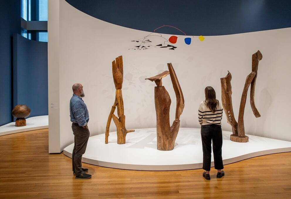 Two people stand in front of wooden sculpture in a gallery.