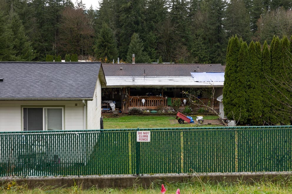 Two houses, one with a porch, and a yard with a wagon are seen behind a green fence.