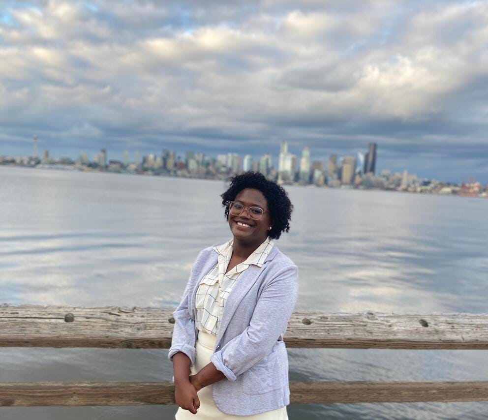 a black woman in a light purple suit stands in front of the puget sound with seattle in the background