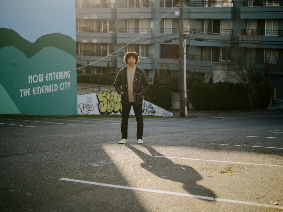 A full body shot of a man standing in a parking lot. He is backlit by the sun and his shadow is visible stretching out in front of him. The wall behind him reads
