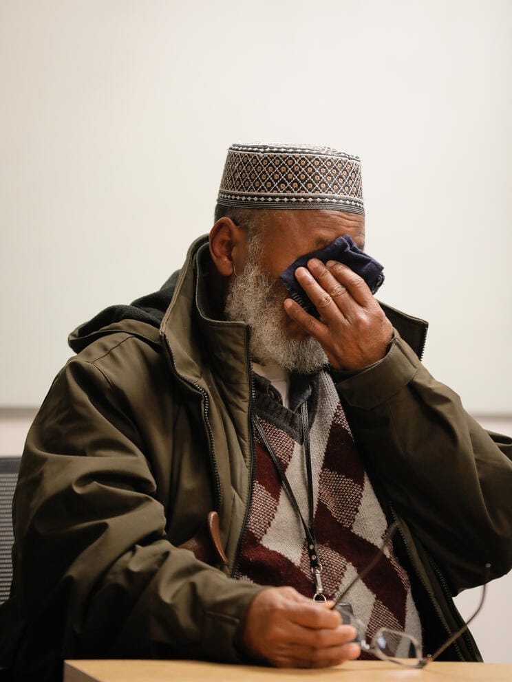 A man sits at a table. His face is hidden by his and a handkerchief he is using to wipe tears away