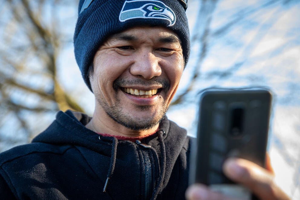 A close up of a man in a Seahawks beanie looking down at his phone and smiling