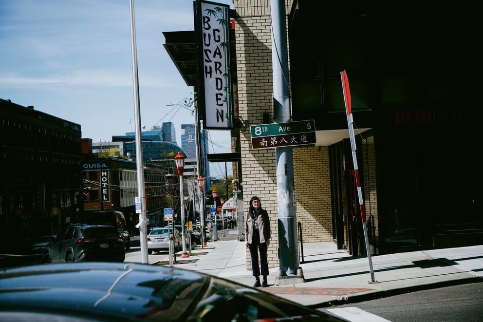 A woman stands on a street corner in the International District. The frame is off kilter and a sign above her head reads Bush Garden.