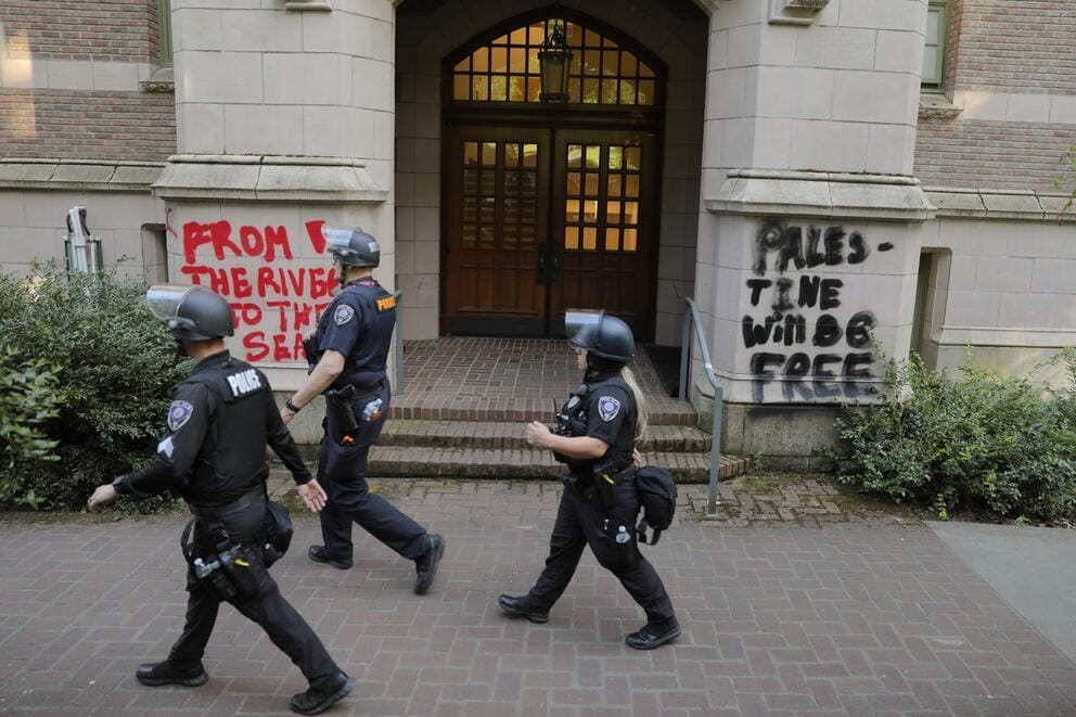 Three uniformed police officers walk past graffiti on the outside of a university building. The left side reads
