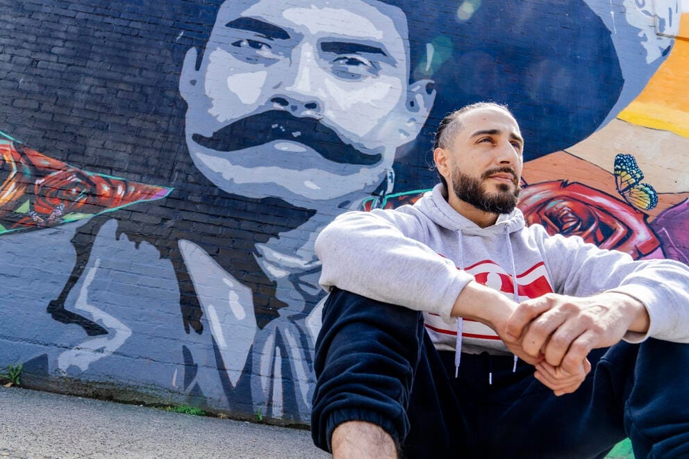 A man sits on the ground in front of a large mural of Emiliano Zapata