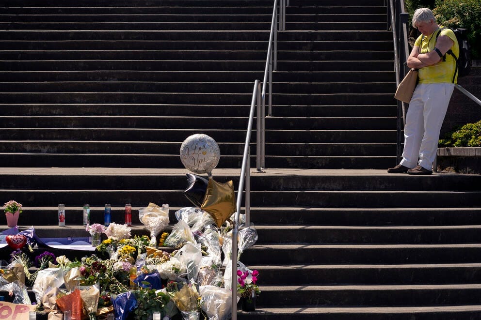 A man stands on a stairway near a memorial filled with flowers, cards, balloons and other trinkets left by mourners