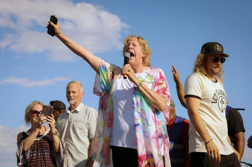 A woman seen from below and surrounded by other people on stage yells into a microphone