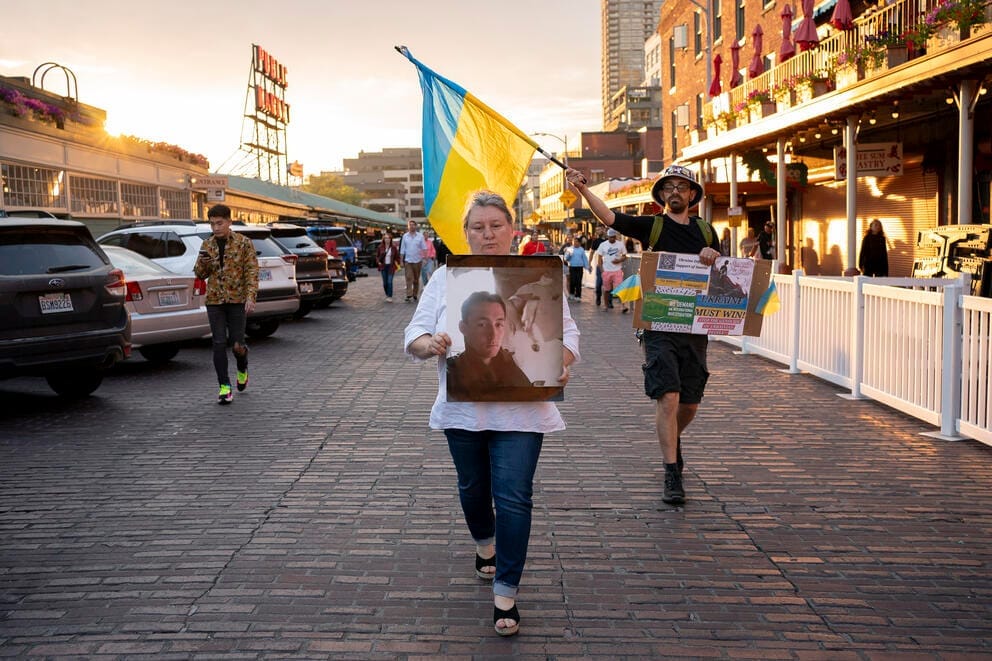 A woman walks down a street holding a photo of a young man, behind her a Ukrainian flag held by another demonstrator