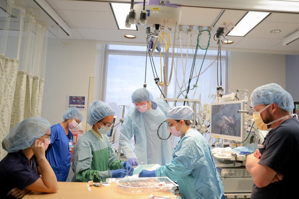 A group of doctors and other medical personnel, all wearing full surgical scrubs, surround a patient on an operating table