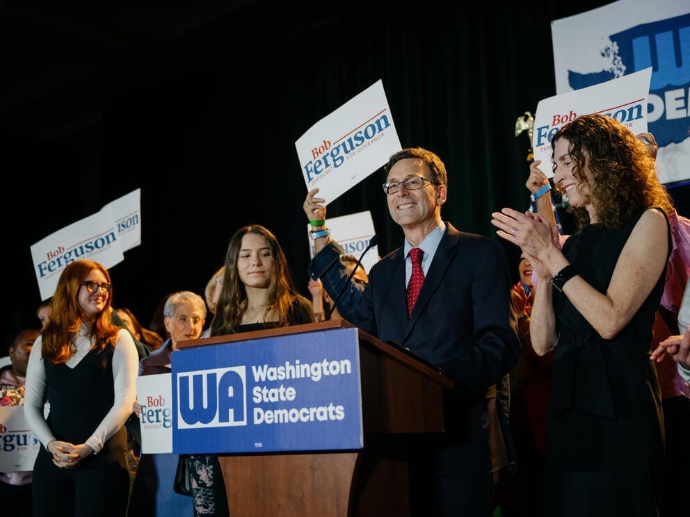 Bob Ferguson smiles behind a podium as family and supporters cheer around him, some holding signs