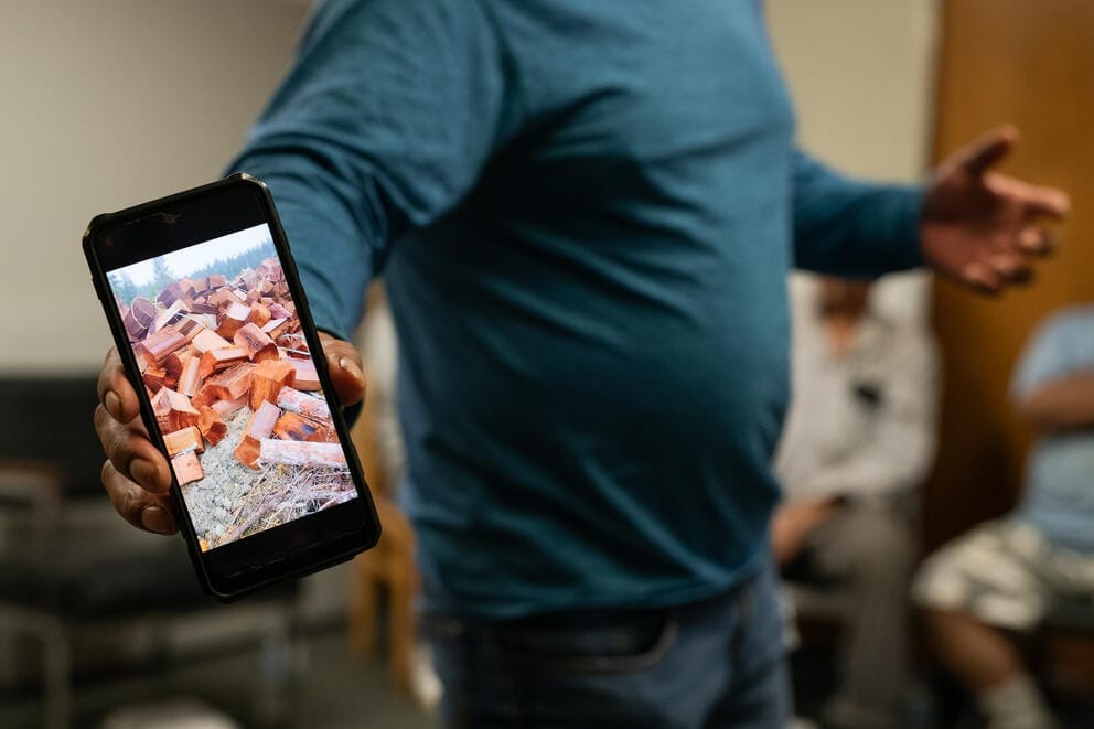 A worker shows his phone to the camera. On it is a photo of the blocks of cedar he's cut to be made into cedar roofing.