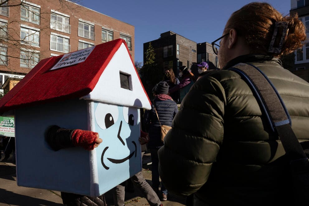 a person dressed in costume shaped like a house. The costume has a red roof, blue sides, and a smiling face